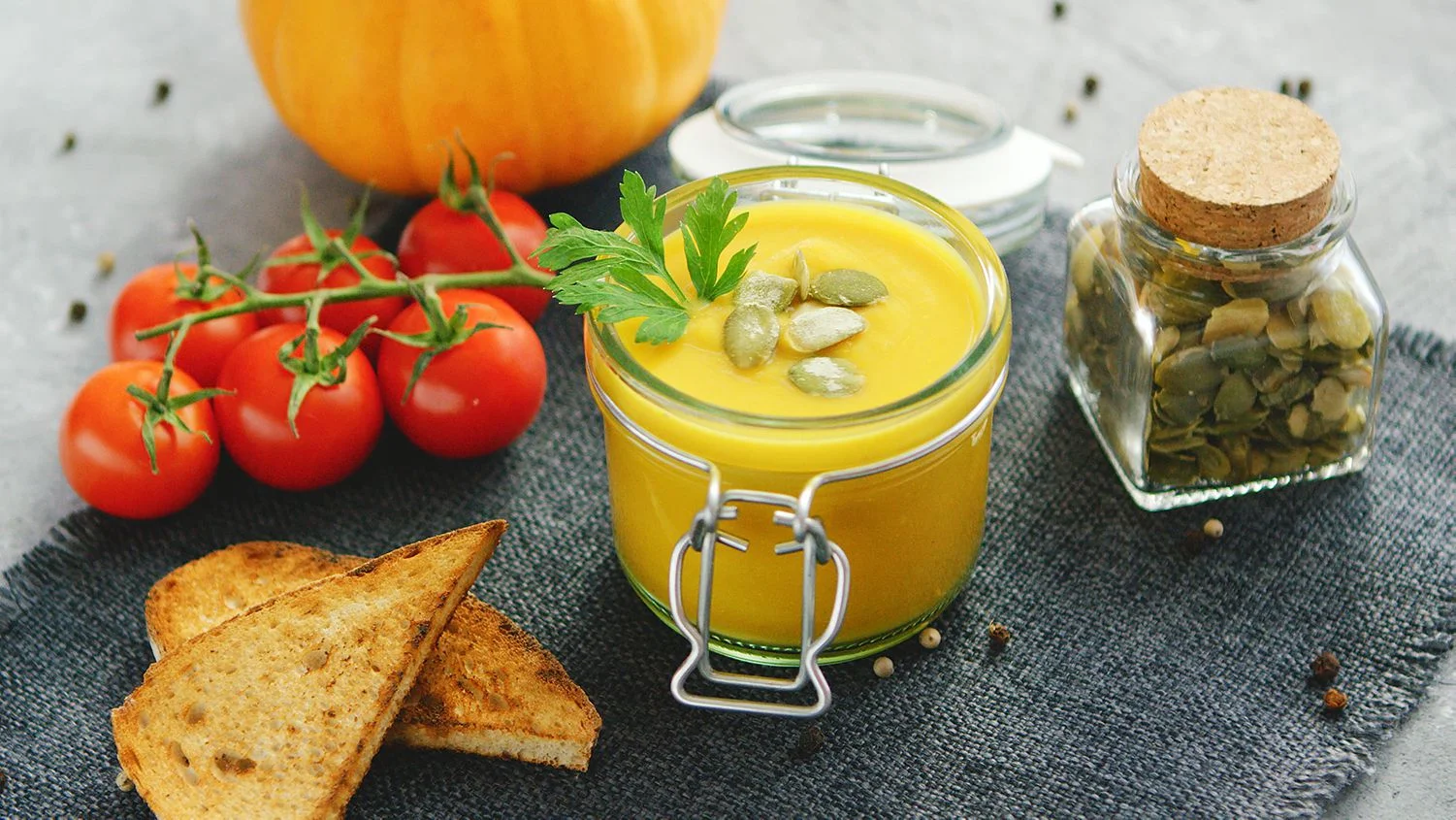 Glasgefäß mit Kürbissuppe, umgeben von frischen Tomaten und Brot auf einer Textilserviette von Tirana